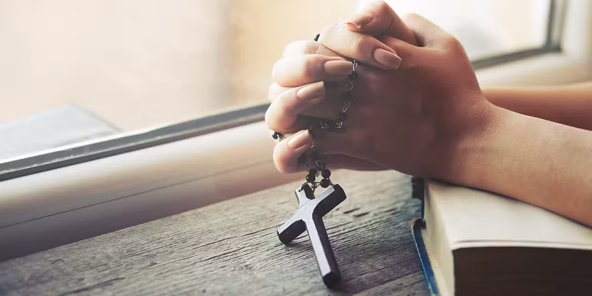 folded hands in prayer holding a rosary with a cross.